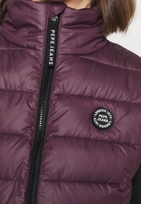 Fialová prošívaná vesta s vysokým límcem, vybavená černým zipem a kulatou nášivkou s logem. Texturovaný prošívaný materiál s hladkým povrchem.