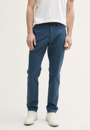 Man in blauwe casual broek, witte sneakers en een wit T-shirt, staand tegen een effen lichte achtergrond.