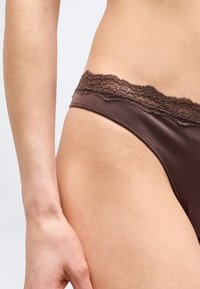 Κοντινό πλάνο στο ισχίο και το χέρι ενός ατόμου που φοράει καφέ εσώρουχο με λεπτομέρειες από δαντέλα, πάνω σε απλό ανοιχτό φόντο.