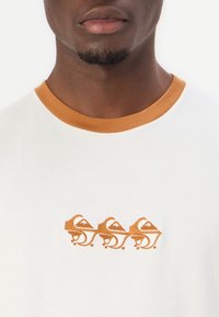 Camiseta blanca con un cuello acanalado naranja. Presenta tres diseños de montañas bordados en naranja en el pecho. Textura suave.
