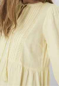 Ljusgul klänning med texturerad tyg, veckad design och spetsdetaljer vid halsringningen, avslutad med en tofs.