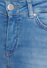 Jeans de denim azul claro com uma textura suave, apresentando costura em contraste, um fecho de botão metálico e um bolso frontal.