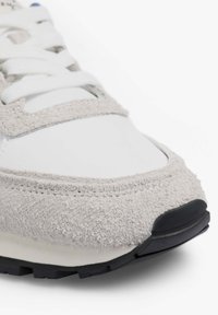 Zapatillas con una parte superior blanca lisa y detalles en gamuza gris, puntera redonda y suela de caucho negra. Los cordones son blancos y las costuras son visibles.