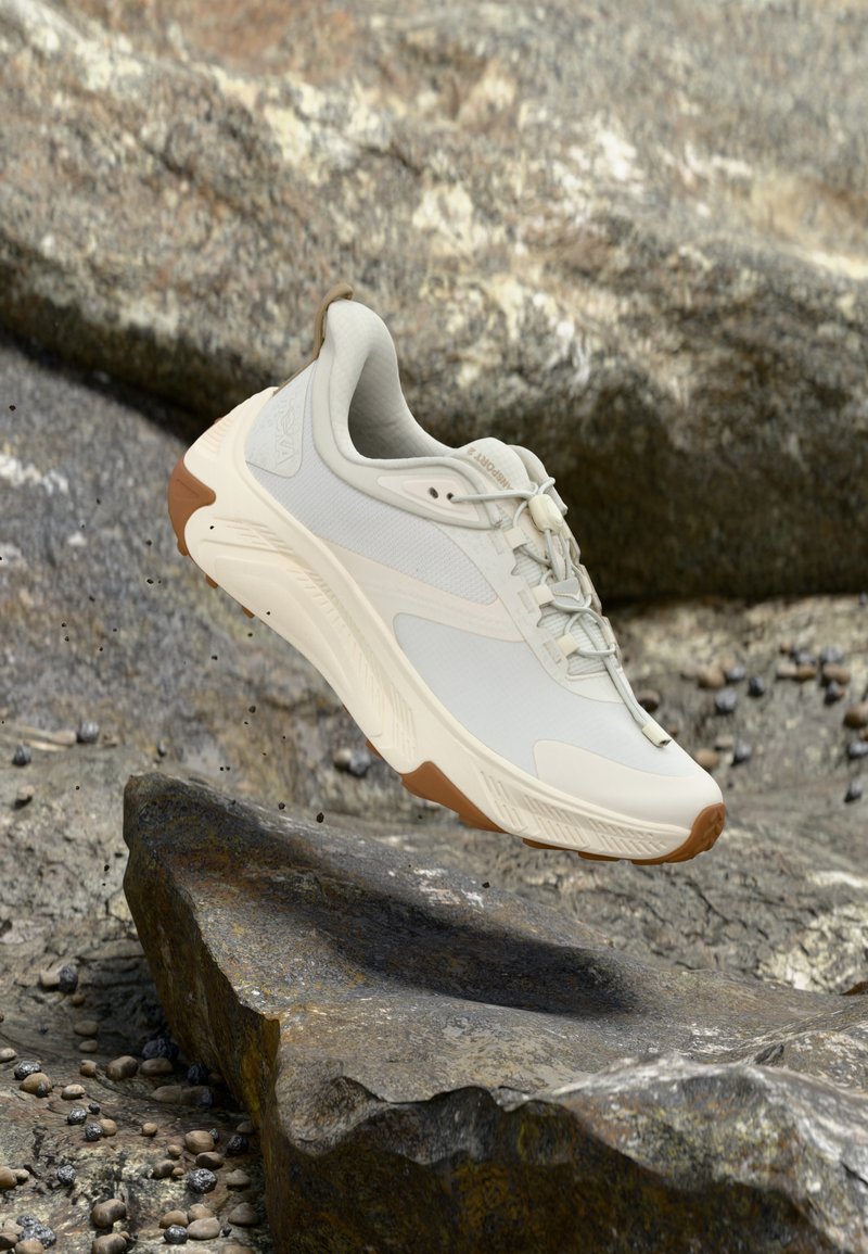 Μπεζ και λευκό αθλητικό παπούτσι με ταμπά σόλα που αιωρείται πάνω από βραχώδες έδαφος με μικρές διασκορπισμένες πέτρες.