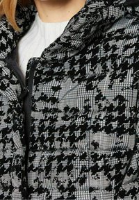 Czarna i biała kurtka puchowa w kratę typu jodełka, o błyszczącej fakturze, z zamkiem błyskawicznym i kapturem zapinanym na zatrzaski.