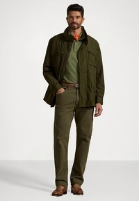 Olive green outer jacket with multiple pockets, worn over a lighter green polo. Dark olive trousers and brown shoes complete the outfit.