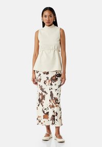 Cremfarbener ärmelloser Top mit hohem Kragen und Peplum-Detail, kombiniert mit einem langen Rock mit einem braun-schwarzen gepunkteten Muster. Weiße Ballerinas.