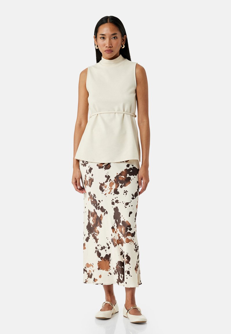 Top sin mangas color crema con cuello alto y detalle peplum, combinado con una falda larga que presenta un patrón de manchas en tonos marrón y negro. Bailarinas blancas.