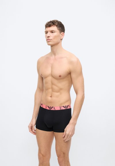 Μυώδης άνδρας να στέκεται χωρίς μπλούζα, φορώντας μαύρα μποξέρ με ροζ μέση που φέρει ένα μαύρο λογότυπο και γράμματα σε απλό φόντο.