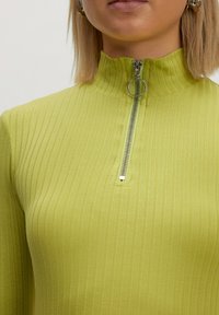 Grünes, geripptes Quarter-Zip-Oberteil mit hohem Kragen, ausgestattet mit einem silbernen Reißverschluss und einem Ring-Zip-Zieher. Weiche Textur mit taillierter Passform.