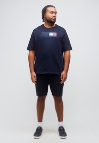 Άνδρας με πλεγμένα μαλλιά, που φοράει μπλε μπλουζάκι Tommy Hilfiger, ταιριαστά σορτς, λευκές κάλτσες και μαύρα αθλητικά παπούτσια, στέκεται μπροστά σε απλό φόντο.