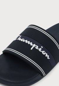 Σαγιονάρα σκούρου μπλε χρώματος με υφασμένο λουρί που διαθέτει λευκό ανάγλυφο λογότυπο "Champion" και δύο λευκές οριζόντιες ρίγες.
