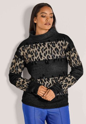 Vrouw in een zwart en bruin luipaardprint coltrui met blauwe broek, handen gekruist voor zich, kijkend opzij.