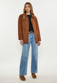 Giubbotto in suede marrone con ampio colletto, chiusura a bottoni e tasche a patta, abbinato a jeans a gamba larga azzurri e stivali alla caviglia marroni.