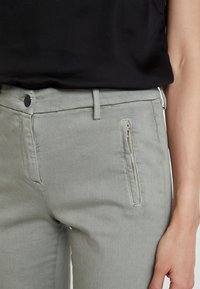 Detailní záběr pasu osoby v šedých kalhotách s bočním zipovým kapsou, spárovaných s černým topem. Látka vypadá strukturovaně.