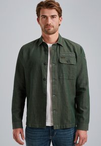 Chemise boutonnée vert foncé en tissu texturé, avec une poche poitrine à patte et un col standard. Portée par-dessus un t-shirt blanc.
