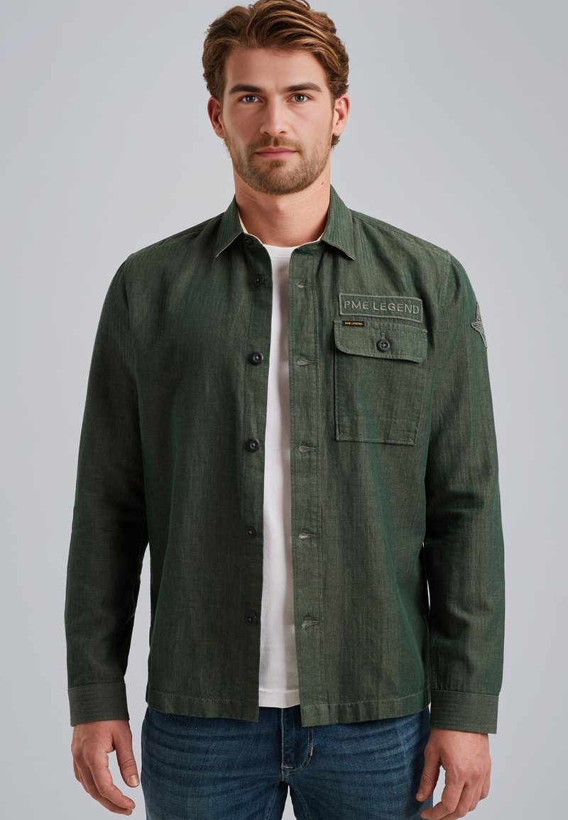 Chemise boutonnée vert foncé en tissu texturé, avec une poche poitrine à patte et un col standard. Portée par-dessus un t-shirt blanc.