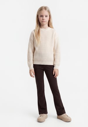 Ung pige med langt blondt hår iført en cremefarvet strikket sweater, mørkebrune bukser og beige platformsneakers, stående mod en hvid baggrund.