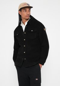 Schwarze Cordjacke mit zwei Brusttaschen, Druckknöpfen und einem gerippten Textur, kombiniert mit einer beigen Mütze mit grafischem Design.