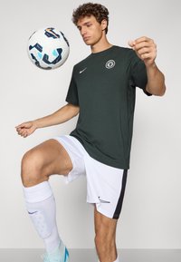 Donkergroen T-shirt met ronde hals en een logo, gecombineerd met witte shorts en schoenen met blauwe accenten. Een voetbal hangt in de lucht.