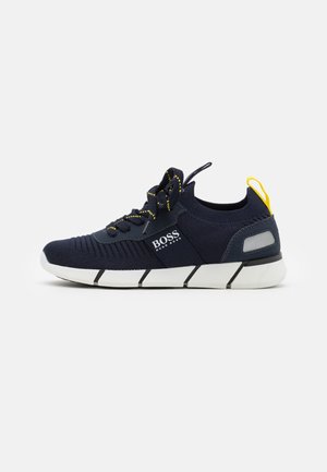 Granatowy dzianinowy sneaker z białą segmentowaną podeszwą, czarno-żółtymi sznurowadłami, żółtą pętelką na pięcie i logo BOSS Hugo Boss po boku.