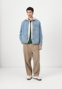 Light blue denim jacket with a shirt collar, two front pockets, layered over a green shirt and beige wide-leg trousers, paired with white sneakers.