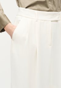 Άτομο που φοράει λευκό παντελόνι με ψηλή μέση και φαρδιά μπατζάκια με πλαϊνές τσέπες, το χέρι στηρίζεται μέσα σε μία από τις τσέπες, συνδυασμένο με μία μπεζ μακρυμάνικη μπλούζα.