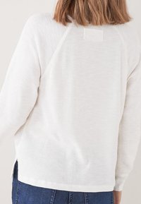Personne portant un haut long à manches longues texturé blanc avec des manches raglan et un jean bleu, vu de dos sur un fond uni.