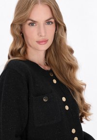 Pulover negru cu guler rotund, având un buzunar texturat și butoane aurii pe față, cu o textură moale și ușor pufoasă.