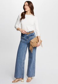 Top bianco a maniche lunghe, jeans blu a vita alta e a gamba larga, borsa a tracolla in suede color sabbia con frange, e sandali con tacco a blocco in oro.