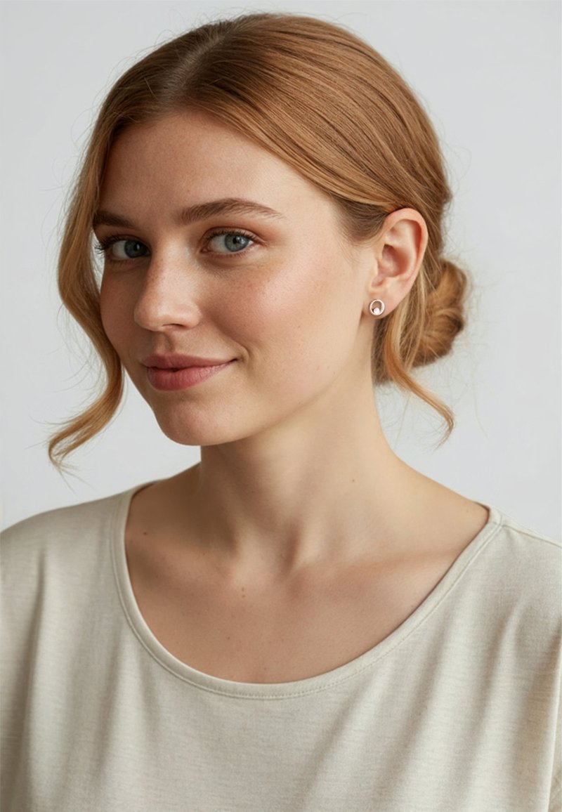 Ljusbeige t-shirt med bred halsringning, mjuk textur och rund form. Modellen har ljusbrunt hår som är uppsatt i en knut och bär små, runda örhängen.