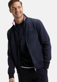 Veste bleu marine zippée avec un col intérieur rose, dotée d'un logo sur la poitrine, de poches latérales et de poignets côtelés. Matière douce, coupe athlétique.