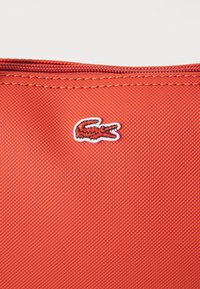 Röd tygväska med en strukturerad yta, med en broderad vit och svart Lacoste-krokodil-logotyp nära dragkedjan.