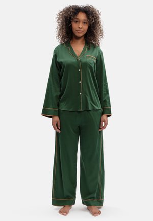 Vrouw staat blootsvoets, draagt een donkergroene satijnen pyjama met bruine bies, een blouse met knopen en losse broek tegen een effen achtergrond.