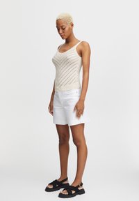 Top tank in maglia beige con un motivo a zigzag, abbinato a pantaloni corti bianchi e sandali neri con cinturini, caratterizzati da una suola spessa.