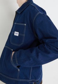 Persona vestida con una chaqueta de denim azul oscuro de Lee, con un bolsillo frontal visible y costuras en contraste blancas, con la mano en el bolsillo lateral, frente a un fondo liso.