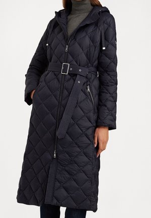 Frau trägt einen langen schwarzen Steppmantel mit Kapuze, tailliertem Gürtel, Reißverschluss und seitlichen Reißverschlusstaschen über einem grauen Rollkragenpullover.