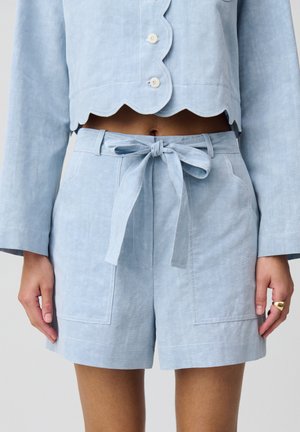Pantalones cortos de lino azul claro con un ajuste relajado, que cuentan con una cintura anudada, bolsillos delanteros y detalles de costura. Textura suave y diseño a medida.