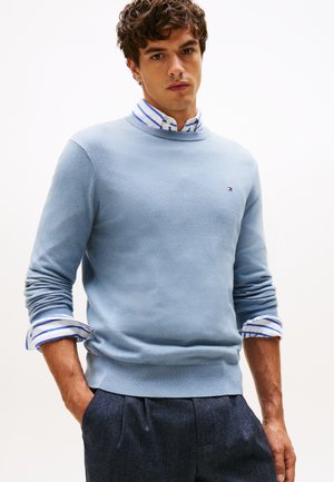 Young man wearing a light blue sweater over a white shirt with blue stripes and dark trousers, standing with hands in pockets.