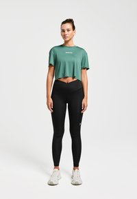 T-shirt vert court à manches courtes avec logo blanc, associé à un legging noir taille haute. Le mannequin porte des baskets blanches.