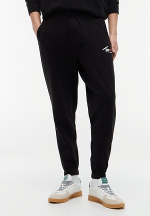 Osoba nosiaca čierne teplákové nohavice s postrannými vreckami a biele tenisky so svetlohnedou podrážkou na bielom pozadí.