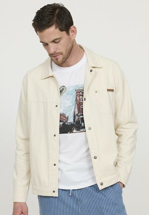 Veste crème claire, boutonnée avec une poche poitrine, associée à un t-shirt blanc avec un design graphique. Pantalons rayés bleus visibles en dessous.