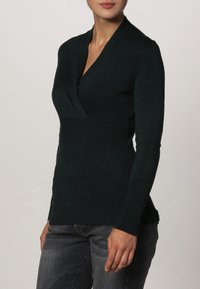 Skovgrøn indpakningsbluse med lange ærmer, ribbet overflade og dyb V-hals, parret med mørkegrå jeans.