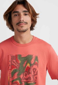 Camiseta de color coral con cuello redondo; presenta un diseño gráfico en verde, rojo y negro que ilustra líneas de contorno y texturas en la parte frontal.