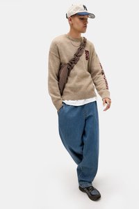 Beige strikket genser med burgunder detaljer, avslappede blå denim-bukser og svart fottøy. Iført en beige og blå cap, og en brun skulderveske.