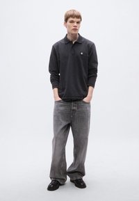 Σκούρα γκρι μπλούζα πόλο με μακριά μανίκια και μικρό λογότυπο, σε συνδυασμό με ανοιχτό γκρι τζιν φαρδιάς γραμμής και μαύρα παπούτσια, δημιουργώντας ένα χαλαρό, καθημερινό στιλ.