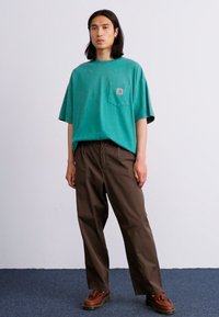 Tricou din bumbac turcoaz cu un buzunar pe piept, asortat cu pantaloni maro lejeri. Modelul stă pe un fundal de carpetă albastră.