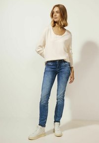 Suéter de punto rosa claro con escote en V y detalles de encaje, combinado con jeans ajustados azules y zapatillas altas blancas.