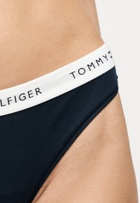 Κοντινό πλάνο ατόμου που φοράει σκούρο μπλε εσώρουχα με λευκή μέση που φέρει το λογότυπο "Tommy Hilfiger" στο ύψος του ισχίου.