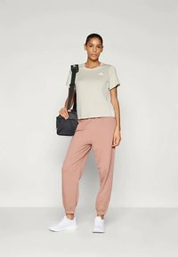 Mulher em pé a usar uma t-shirt Adidas cinza claro, calças de fato de treino cor-de-rosa, ténis brancos e a segurar uma mala à tiracolo preta, contra um fundo simples.